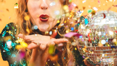girl in green sequin dress blowing confetti towards the camera holding silver disco ball